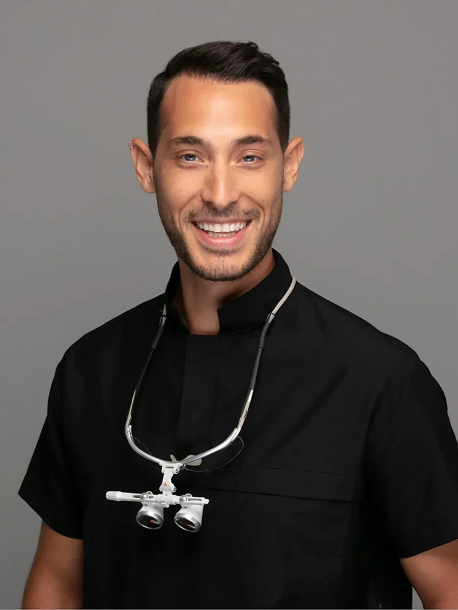 Dt. Doğa Erginoğlu smiling, wearing black scrubs with dental loupes around his neck